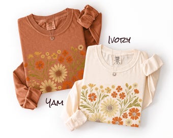 May include: Two long-sleeve shirts, one in yam and one in ivory, each featuring a graphic print of orange, yellow, and green wildflowers across the lower front. Both shirts have a crew neckline and a Comfort Colors brand label at the inner collar.
