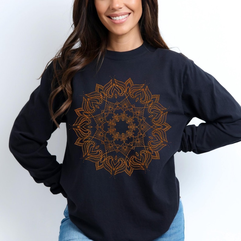 May include: Black long-sleeve shirt featuring a detailed, golden mandala design on the front. The intricate pattern is centered and symmetrical, with heart-shaped elements. The shirt is a solid color, with a crew neck and long sleeves.