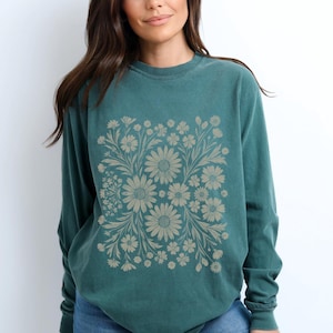 May include: Teal-colored long-sleeve sweatshirt featuring a floral design. The design is a square arrangement of beige daisies and other flowers with leaves. The sweatshirt is made of a soft material and has a crew neck.
