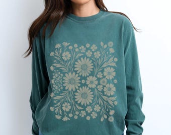 May include: Teal-colored long-sleeve sweatshirt featuring a floral design. The design is a square arrangement of beige daisies and other flowers with leaves. The sweatshirt is made of a soft material and has a crew neck.