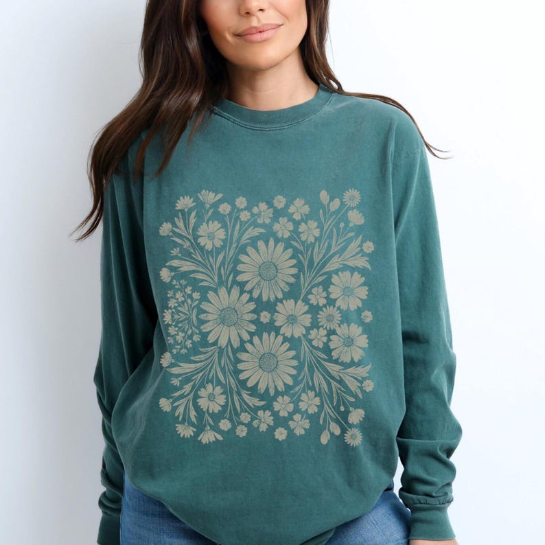 May include: Teal-colored long-sleeve sweatshirt featuring a floral design. The design is a square arrangement of beige daisies and other flowers with leaves. The sweatshirt is made of a soft material and has a crew neck.