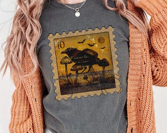 May include: A charcoal gray t-shirt featuring a rectangular graphic designed to look like a postage stamp. The graphic displays a yellow background with dark silhouettes of mushrooms, bats, and a moon, along with the number 10 and text.