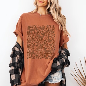 May include: Rust-colored t-shirt featuring a brown woodland animal and floral design. The shirt has short sleeves and a relaxed fit. A black and white plaid shirt is draped over the shoulders.