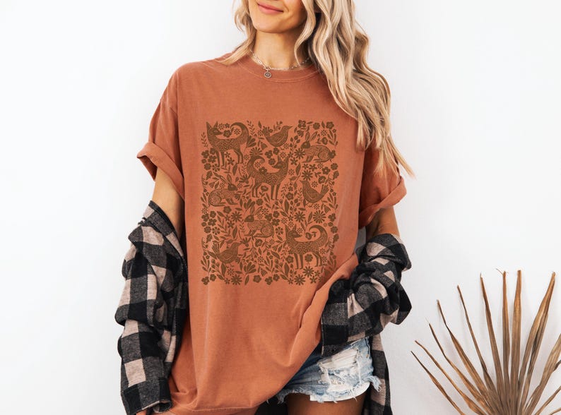 May include: Rust-colored t-shirt featuring a brown woodland animal and floral design. The shirt has short sleeves and a relaxed fit. A black and white plaid shirt is draped over the shoulders.