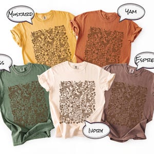 May include: Five different colored t-shirts: mustard, yam, espresso, ivory, and moss. Each shirt features a brown floral and animal pattern. The shirts are short-sleeved and have a relaxed fit.