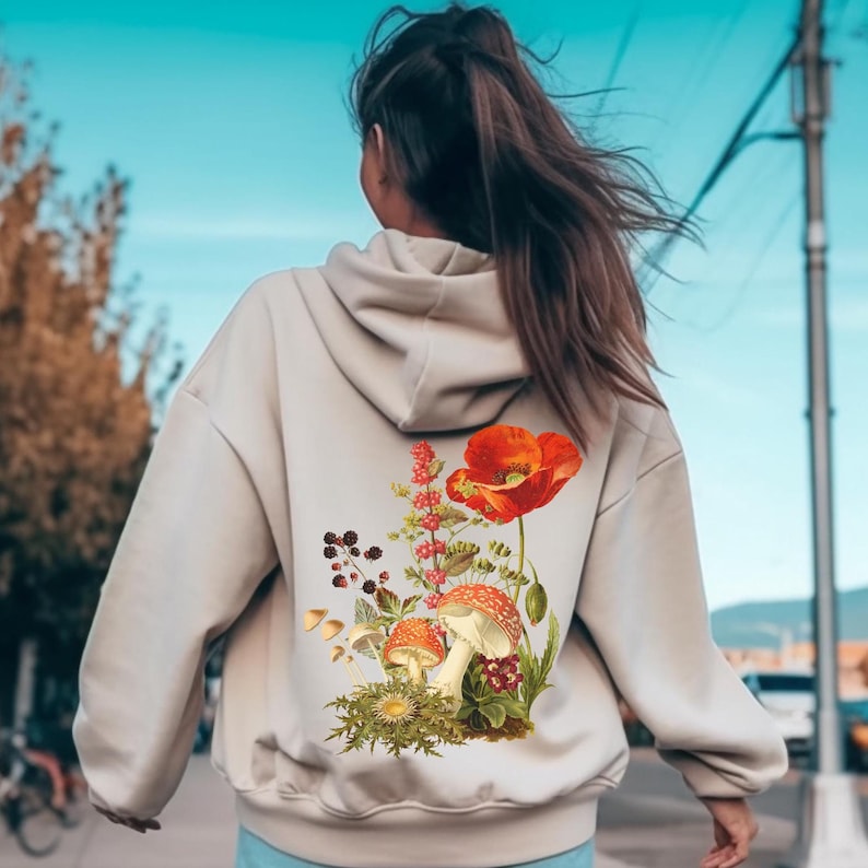 May include: A beige hoodie with a floral design featuring a red poppy, mushrooms, and green leaves.
