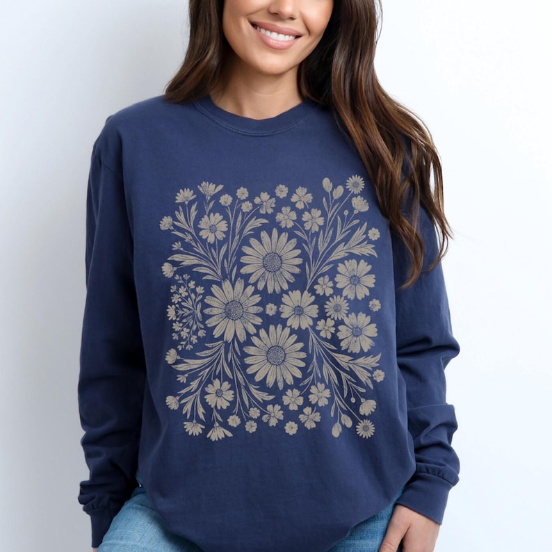 May include: Navy blue long-sleeve sweatshirt featuring a floral design in shades of tan and beige. The floral pattern is centered on the front of the sweatshirt, creating a symmetrical design. The sweatshirt has a crew neck and long sleeves.