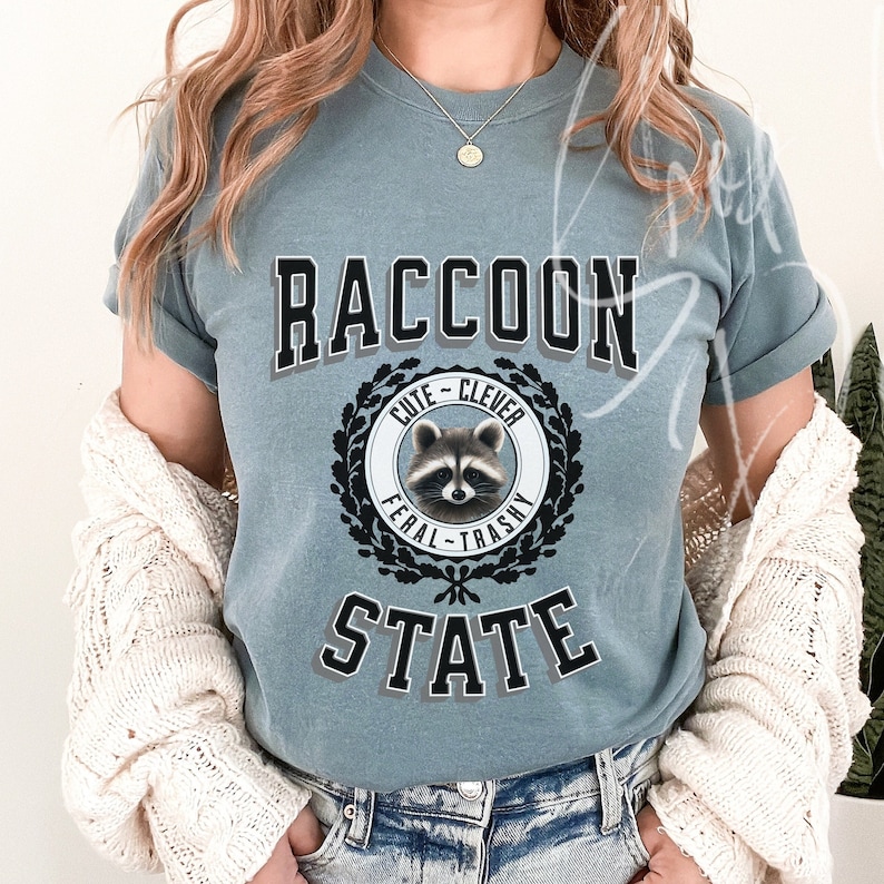 May include: A blue t-shirt with a graphic of a raccoon in a circle with the words "Cute - Clever" above the raccoon and "Feral - Trashy" below the raccoon. The words "Raccoon State" are printed below the circle.