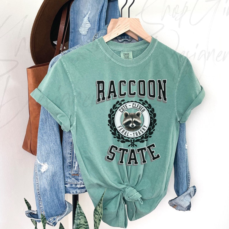 May include: A teal blue t-shirt with a black and white graphic of a raccoon with the text "Raccoon State" and the words "Cute - Clever - Feral - Trashy" in a wreath around the raccoon.