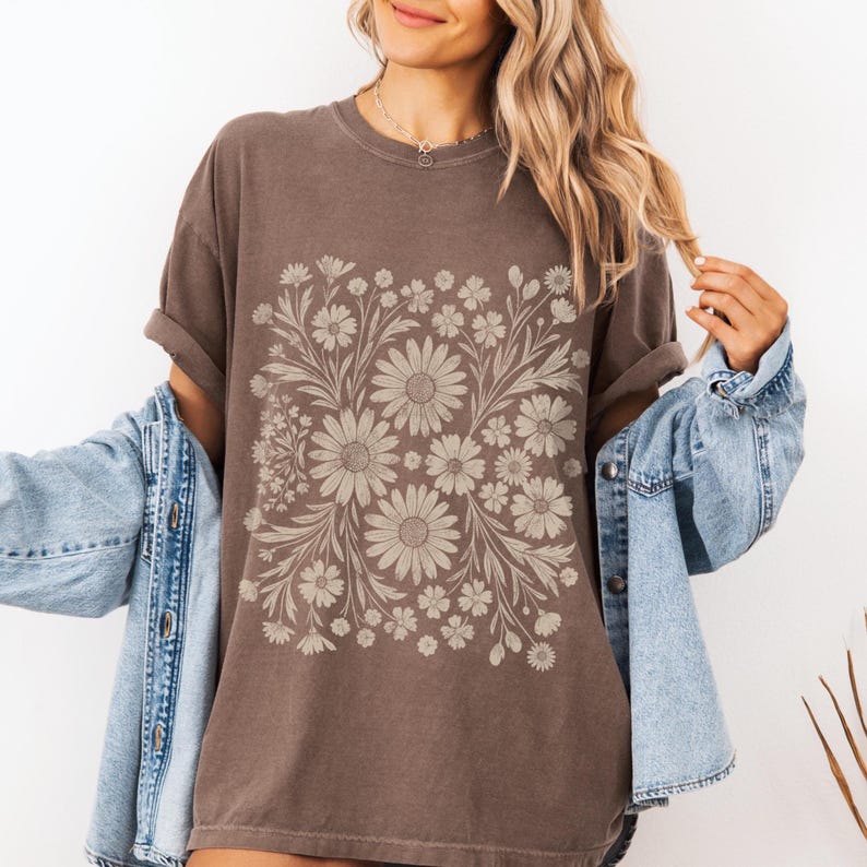Pode incluir: Uma camiseta marrom com um design floral com margaridas e outras flores em um tom marrom mais claro. A camiseta tem mangas curtas e um corte folgado. A modelo est&aacute; usando uma jaqueta jeans azul.