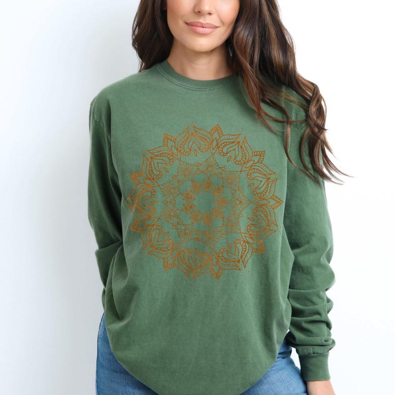 May include: A green long-sleeve shirt featuring a detailed, golden mandala design on the front. The shirt has a relaxed fit and is made of a soft material. The design is centered on the chest.