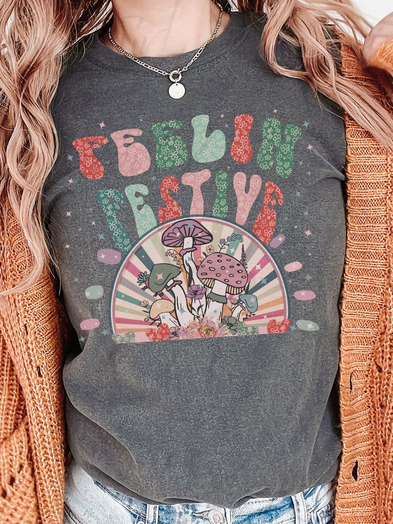 Peut inclure: Un t-shirt gris fonc&eacute; avec le texte "Feeling Festive" dans une police r&eacute;tro avec des fleurs color&eacute;es. Le design comprend &eacute;galement un arc-en-ciel et des champignons.