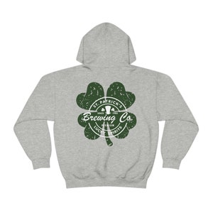 May include: A heather gray hoodie with a green shamrock design. The shamrock features the text "St. Patrick's Brewing Co. Since 1982" and "Premium Lucky Spirits". The hoodie has a drawstring hood and a kangaroo pocket.