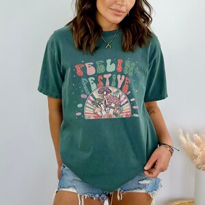 Peut inclure: Un t-shirt vert sarcelle avec un motif de champignons et le texte "Feelin' Festive" dans une police r&eacute;tro.