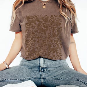 May include: A brown t-shirt featuring a detailed pattern of woodland creatures and floral elements. The design includes deer, birds, foxes, and rabbits, creating a nature-inspired aesthetic. The shirt is paired with blue jeans.