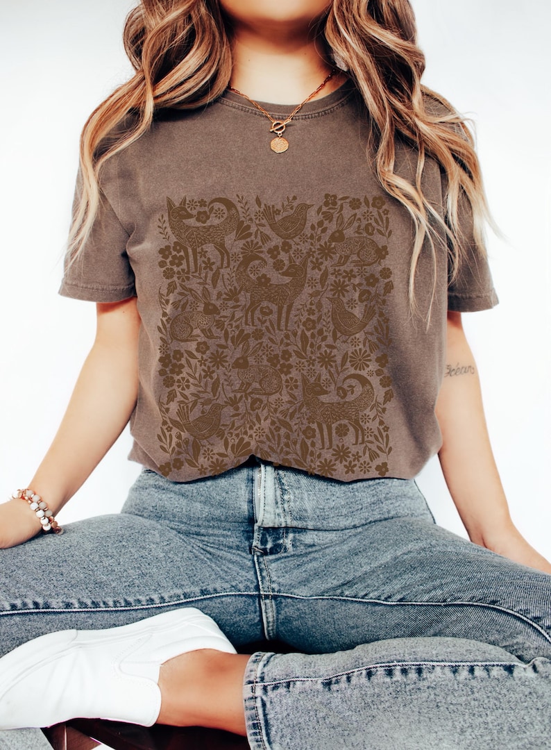 May include: A brown t-shirt featuring a detailed pattern of woodland creatures and floral elements. The design includes deer, birds, foxes, and rabbits, creating a nature-inspired aesthetic. The shirt is paired with blue jeans.