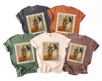 May include: Five short-sleeve crew neck t-shirts in charcoal, orange, green, cream, and brown. Each features a graphic of a postage stamp with a witch holding a broom and pumpkin, a black cat, the number 12, and a circular postmark.