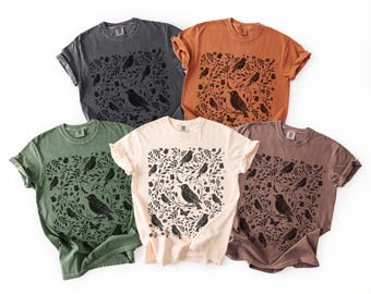 May include: Five short-sleeved crew neck t-shirts in charcoal, burnt orange, sage green, cream, and mauve. Each shirt features a central black graphic print of various birds, butterflies, and leafy botanical branches arranged in a dense pattern.