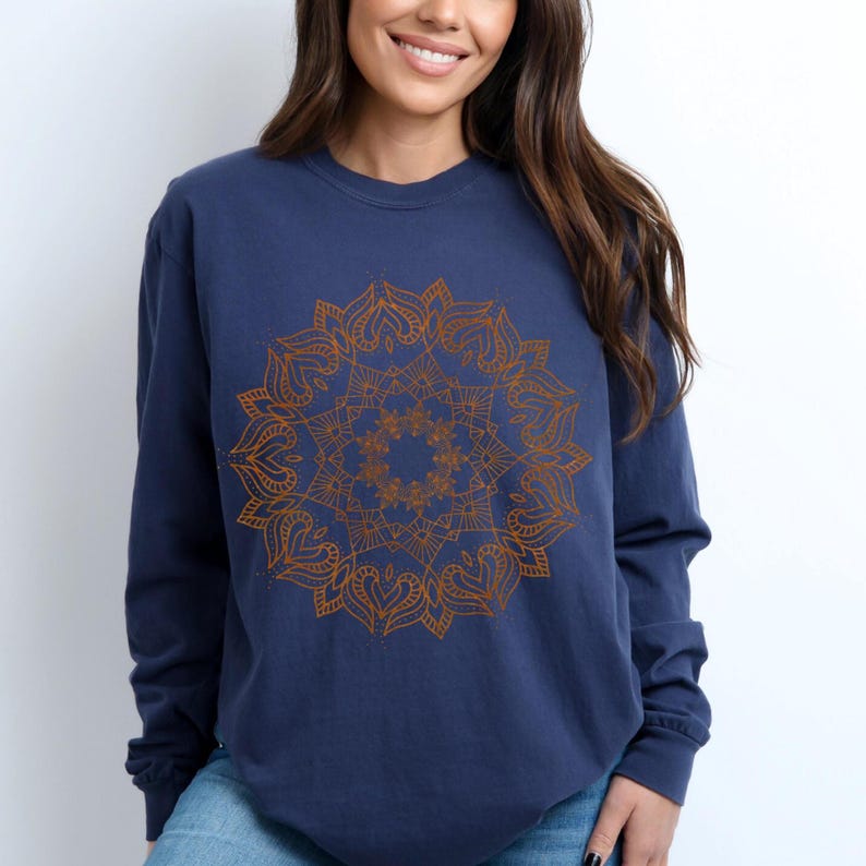 May include: Navy blue long-sleeve sweatshirt featuring a detailed, copper-colored mandala design on the front. The mandala is a symmetrical, circular pattern with intricate floral and heart-shaped elements. The sweatshirt is made of a soft, comfortable material.