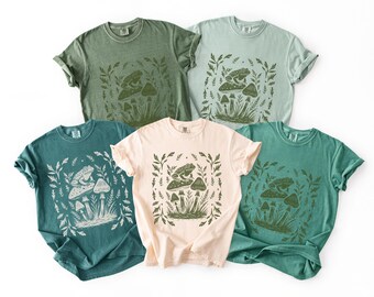 May include: Five short-sleeve crew neck t-shirts in various shades of green and light pink. Each shirt features a central graphic print of a frog sitting on a mushroom surrounded by smaller mushrooms and leafy foliage in a rectangular frame.