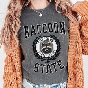May include: A gray t-shirt with the text "RACCOON STATE" in black block letters. A black and white illustration of a raccoon with a wreath around it says "Cute - Clever - Feral - Trashy".