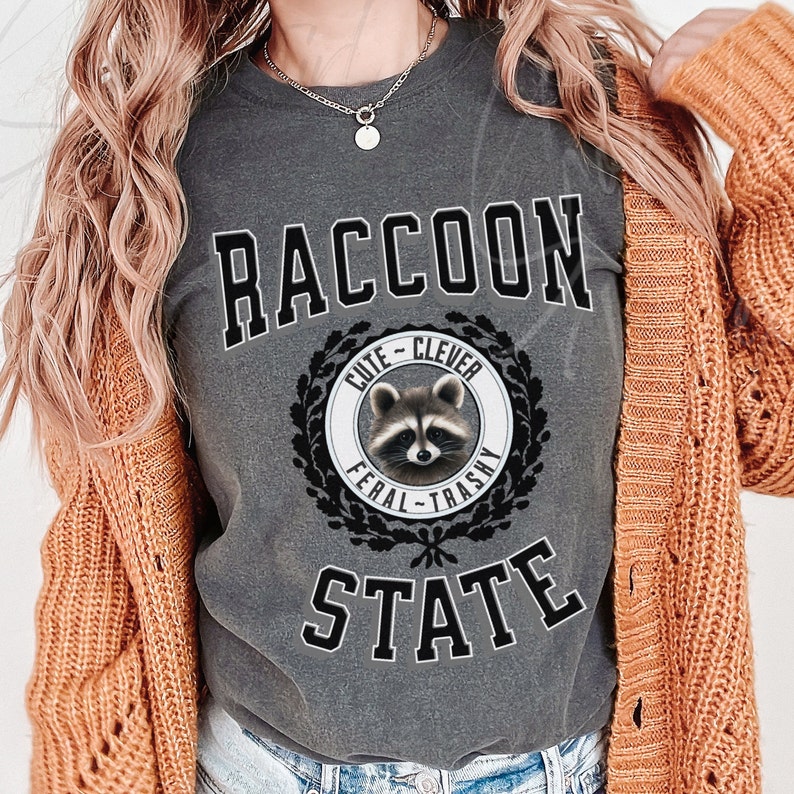 May include: A gray t-shirt with the text "RACCOON STATE" in black block letters. A black and white illustration of a raccoon with a wreath around it says "Cute - Clever - Feral - Trashy".