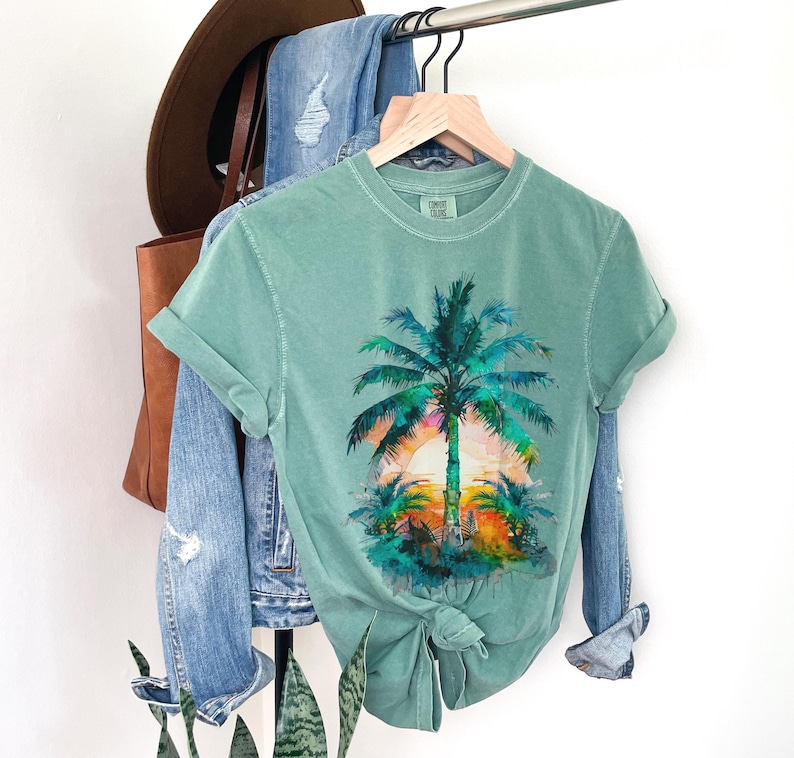 May include: A seafoam green t-shirt featuring a watercolor design of a palm tree at sunset. The shirt is hanging on a wooden hanger, with a pair of blue jeans and a brown hat in the background. The shirt is tied in a knot at the bottom.