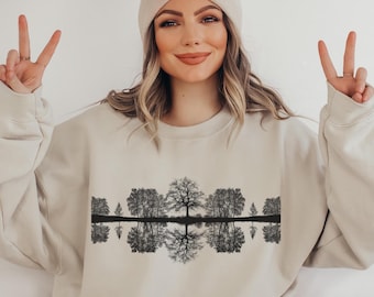 May include: A beige sweatshirt with a black and white graphic print of trees reflected in water. The sweatshirt has a crew neck and long sleeves.