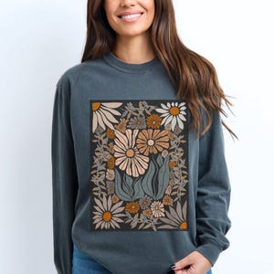May include: A long-sleeved, dark teal sweatshirt featuring a floral design. The graphic includes various flowers in shades of beige, orange, and brown, set against a black background. The design is framed by a border of leaves and vines.