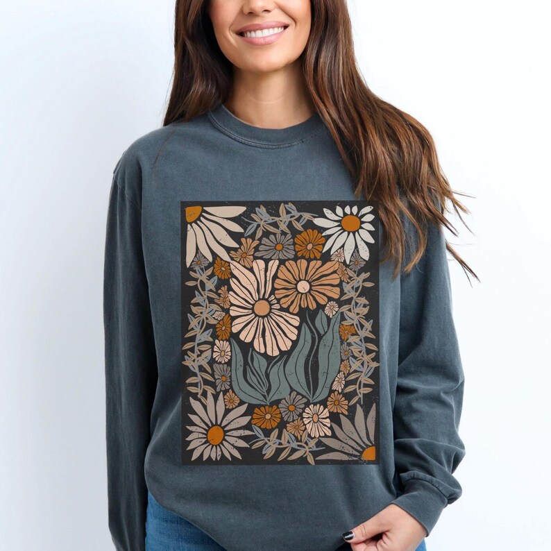 May include: A long-sleeved, dark teal sweatshirt featuring a floral design. The graphic includes various flowers in shades of beige, orange, and brown, set against a black background. The design is framed by a border of leaves and vines.