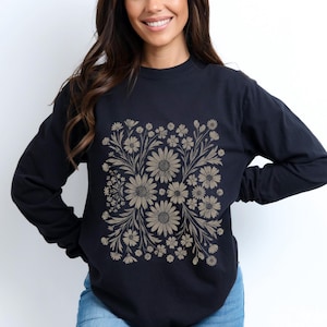 May include: A navy blue long-sleeve shirt featuring a floral design in shades of tan and beige. The design is a square pattern of daisies and other flowers with leaves and stems. The shirt has a crew neck.