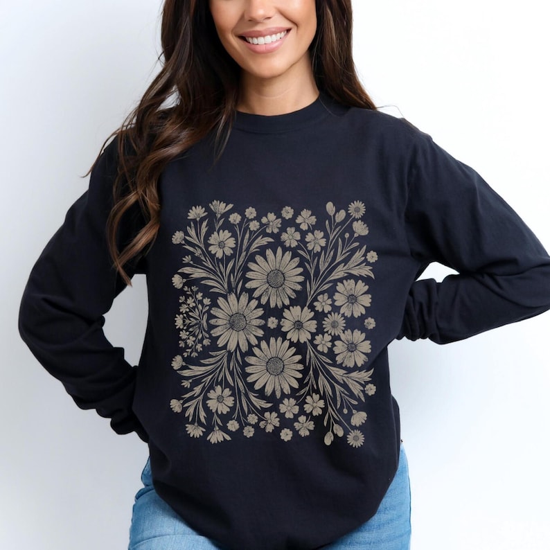 May include: A navy blue long-sleeve shirt featuring a floral design in shades of tan and beige. The design is a square pattern of daisies and other flowers with leaves and stems. The shirt has a crew neck.