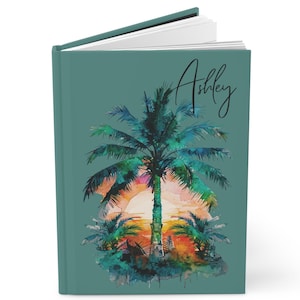 Könnte beinhalten: Ein tealfarbenes Notizbuch mit dem Namen "Ashley" in Schreibschrift. Das Cover zeigt ein Aquarellgemälde einer Palmensilhouette vor einem Sonnenuntergang mit orange, gelben und rosa Farbtönen.