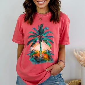 May include: Coral-colored t-shirt featuring a vibrant palm tree graphic with a sunset backdrop. The design showcases a detailed illustration of a palm tree with green fronds and a colorful sky. The shirt is a casual, short-sleeved style.