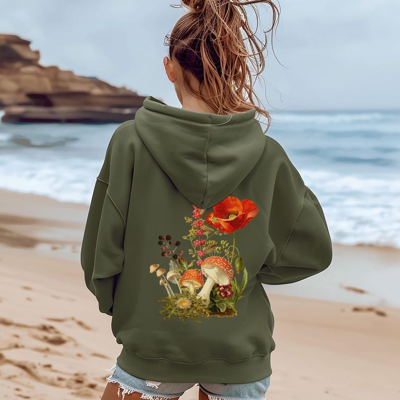 May include: Olive green hooded sweatshirt featuring a nature-inspired back print. The design showcases a vibrant red poppy flower, various mushrooms, and other botanical elements, creating a whimsical, earthy aesthetic. The sweatshirt appears soft and comfortable, suitable for casual wear.