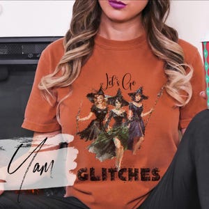Pode incluir: Uma t-shirt de manga curta cor de ferrugem com uma ilustração gráfica de três bruxas com vestidos pretos e chapéus pontiagudos. O texto "Let's Go" está impresso acima das bruxas e a palavra "GLITCHES" está em letras pretas abaixo delas.
