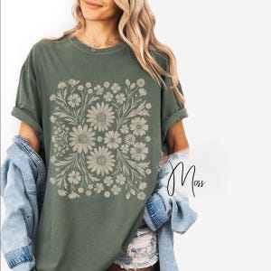 Pode incluir: T-shirt verde-oliva com um design floral com margaridas e outras flores em um arranjo quadrado. O design &eacute; em uma cor neutra, criando um contraste com a cor da camisa. A camisa tem mangas curtas e um corte folgado.
