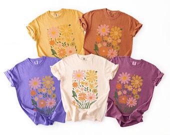 May include: Five short-sleeved crew neck t-shirts in yellow, rust orange, purple, cream, and dark magenta. Each shirt features a rectangular graphic print on the chest displaying a cluster of various flowers in shades of pink, orange, and yellow.