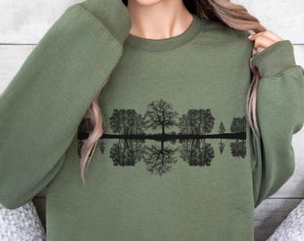 May include: Olive green sweatshirt featuring a black graphic of trees reflected in water. The design is centered on the chest. The sweatshirt has a crew neck and long sleeves. The background is a light wood panel.