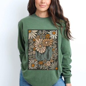 May include: Green long-sleeve shirt featuring a floral design. The design includes large daisies in cream, tan, and orange hues, surrounded by leaves and smaller flowers on a dark background. The shirt is a solid green color.