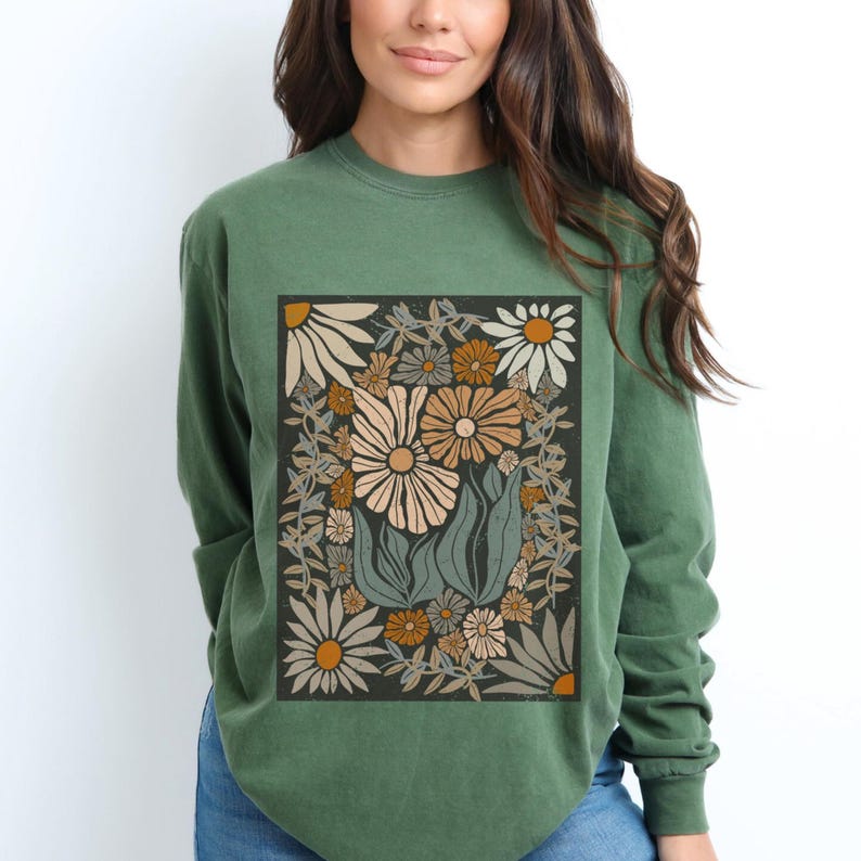 May include: Green long-sleeve shirt featuring a floral design. The design includes large daisies in cream, tan, and orange hues, surrounded by leaves and smaller flowers on a dark background. The shirt is a solid green color.