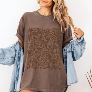 May include: A brown t-shirt featuring a detailed, repeating pattern of deer, birds, and floral elements. The design is in a darker shade of brown, creating a subtle contrast against the shirt's base color. The shirt has short sleeves and a relaxed fit.