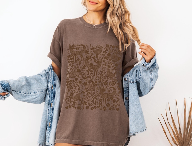 May include: A brown t-shirt featuring a detailed, repeating pattern of deer, birds, and floral elements. The design is in a darker shade of brown, creating a subtle contrast against the shirt's base color. The shirt has short sleeves and a relaxed fit.