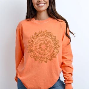 May include: An orange long-sleeve sweatshirt featuring a detailed brown mandala design on the front. The mandala is a symmetrical, circular pattern with intricate floral and heart-shaped elements. The sweatshirt is a solid color and has a relaxed fit.