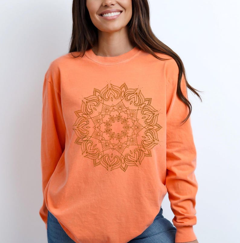 May include: An orange long-sleeve sweatshirt featuring a detailed brown mandala design on the front. The mandala is a symmetrical, circular pattern with intricate floral and heart-shaped elements. The sweatshirt is a solid color and has a relaxed fit.