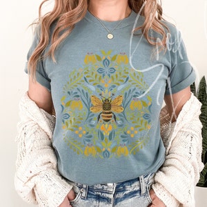 May include: A blue t-shirt with a bee and floral design. The bee is yellow and black and is surrounded by yellow flowers and green leaves.