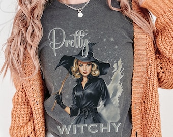 May include: A dark gray t-shirt featuring a graphic of a blonde character wearing a black witch hat and a black dress, holding a wand. The text "Pretty" is printed above the character and "WITCHY" is printed below in a light gray font.