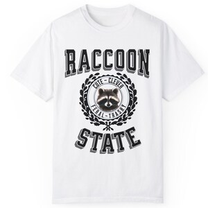 May include: White t-shirt with a black graphic of a raccoon in a circle with the words "Cute - Clever - Feral - Trashy" around it. The words "Raccoon State" are printed below the circle.