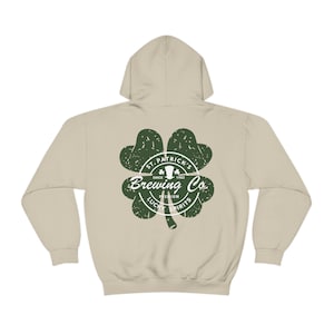 May include: A beige hoodie with a green shamrock design. The shamrock features the text "St. Patrick's Brewing Co. Since 1982" and "Premium Lucky Spirits". The hoodie has a drawstring hood and long sleeves.