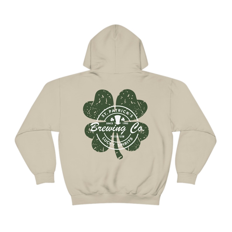 May include: A beige hoodie with a green shamrock design. The shamrock features the text "St. Patrick's Brewing Co. Since 1982" and "Premium Lucky Spirits". The hoodie has a drawstring hood and long sleeves.